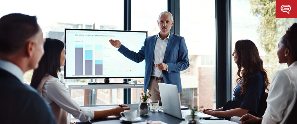 A group of professionals in a conference room is listening to a man giving a presentation. He stands beside a screen displaying charts and graphs from his slide template. The room has large windows with natural light streaming in, and there are plants visible outside.