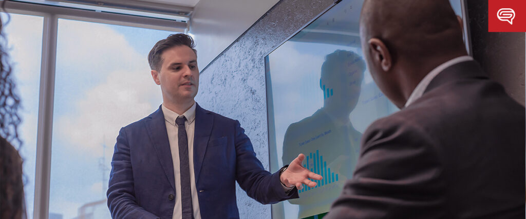 A man in a suit and tie stands and gestures towards a PowerPoint presentation on a large screen, addressing a group of seated individuals in a bright, modern office setting. The screen displays various bar graphs and data charts.