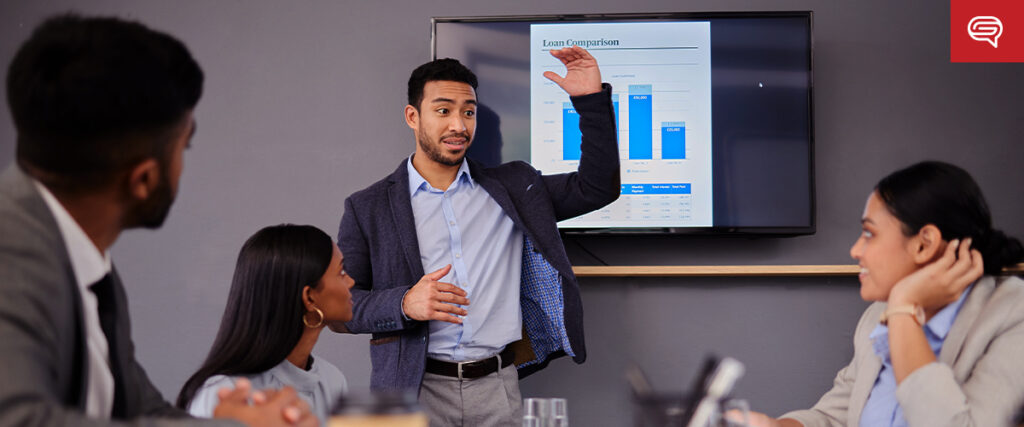 A man in a business suit is standing and gesturing while giving a presentation to three seated colleagues in a conference room. A screen behind him displays a "Loan Comparison" chart. The colleagues appear engaged in the discussion, attentively following the PowerPoint slides.