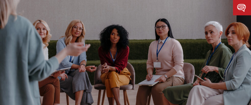 A group of six women sits in a semi-circle, attentively listening to a speaker who stands in the foreground. In this workshop setting, they hold notebooks and pens. The background includes a green cushioned bench, resembling visuals often seen in a pitch deck or PowerPoint presentation.