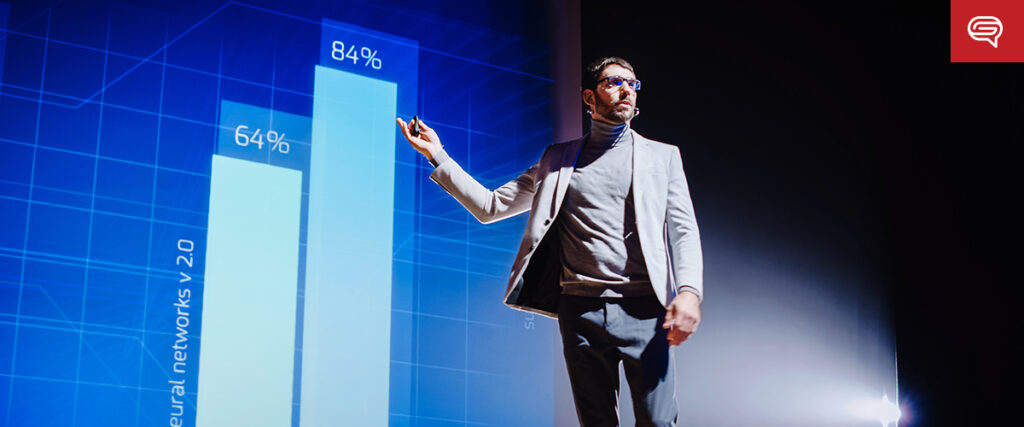A man in a suit and glasses is giving a presentation on stage. He is pointing to a bar graph on a large screen showing percentages of 64% and 84%. The slide has a blue background with a grid pattern, and a speech bubble icon is visible in the top-right corner.
