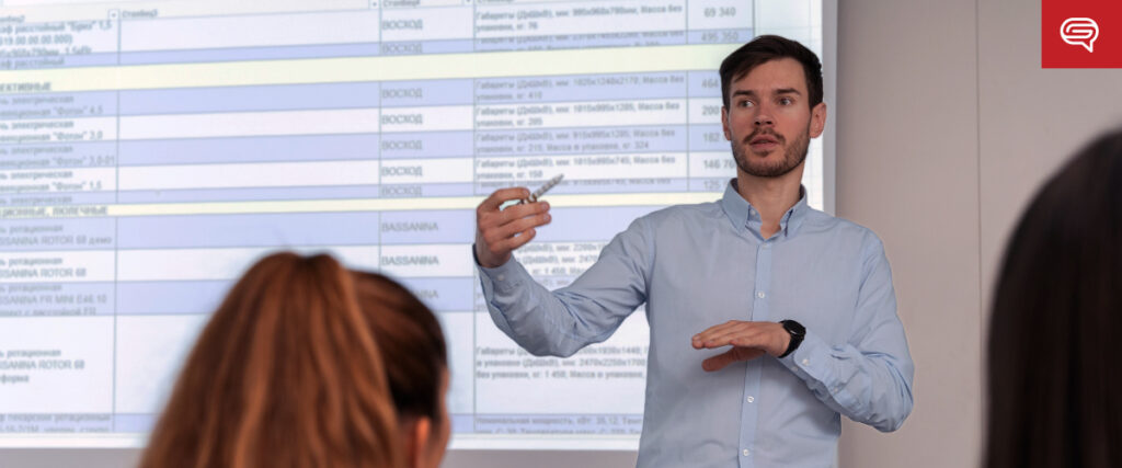 A man in a light blue shirt stands in front of a class, gesturing with his hand while holding a marker. Behind him, a projected presentation displays a pitch deck. Two people are visible in the foreground, facing the instructor.