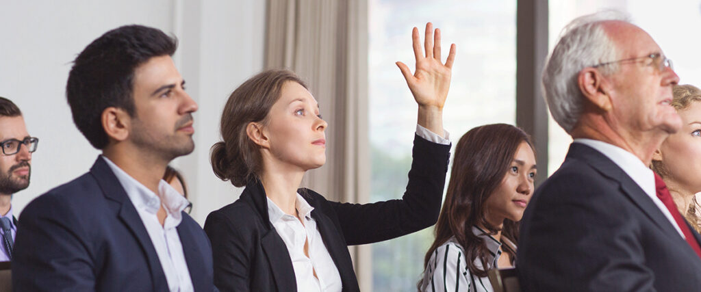 A diverse group of people sit in a seminar room. A woman in the middle raises her hand, indicating she wants to ask a question or participate. The others around her are attentively listening to someone outside the frame as a PowerPoint slide is projected on the screen.