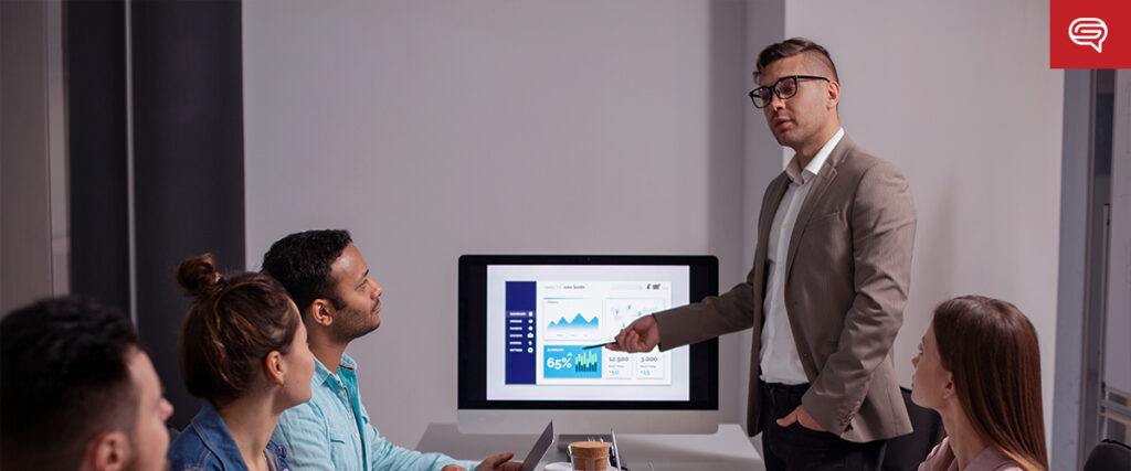 A man in glasses and a suit presents data on a large monitor to a group of four seated individuals in a modern office setting. Using a slide template, the screen shows various charts and graphs. Some of the participants are holding tablets or laptops, observing attentively as if preparing for an important pitch deck.