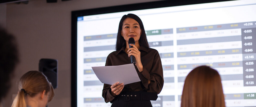 A person stands holding a microphone and a sheet of paper, speaking in front of a large screen displaying a data chart from the pitch deck. The audience, with some people seen from the back, is focused on the speaker.