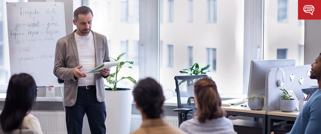 A man in a gray blazer and white shirt is standing and holding documents while giving a presentation to a group of seated individuals in a classroom-like setting. Behind him is a whiteboard with written notes and large windows letting in natural light.