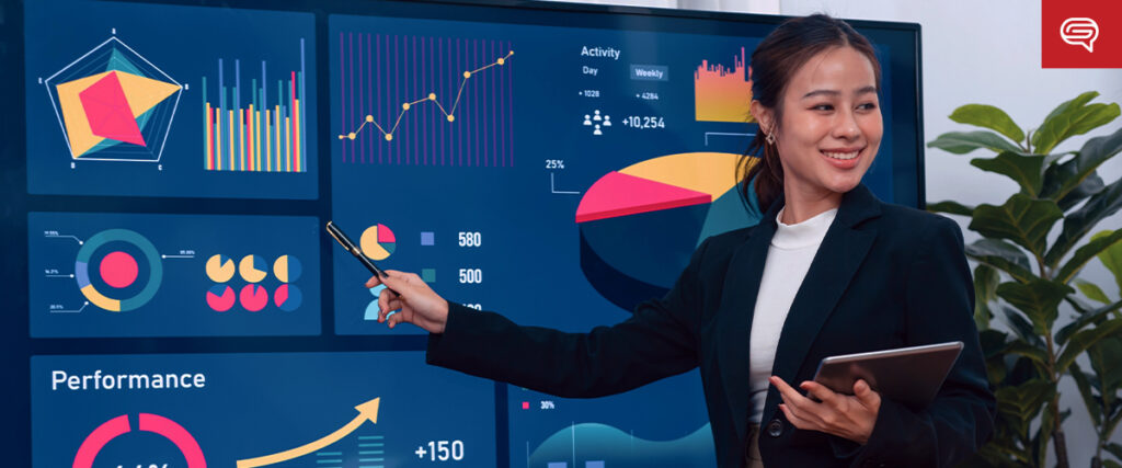 A woman in a business suit stands in front of a large screen displaying various charts and graphs. She is holding a tablet in one hand and pointing to the presentation slide with a stylus. The graphics include bar charts, line graphs, pie charts, and performance metrics.