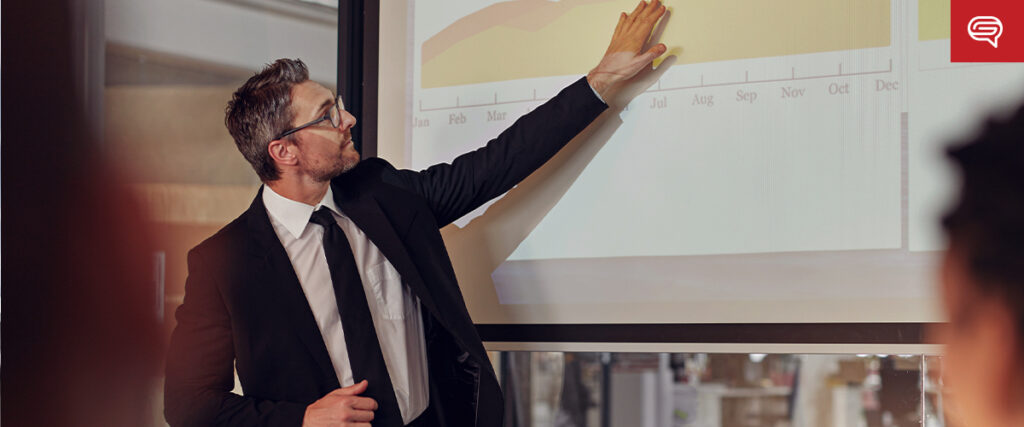 A man in a black suit and glasses points at a line graph projected on a screen during a PowerPoint presentation. The graph depicts data trends across months. The room appears to be a conference or meeting space, with blurred attendees watching intently.