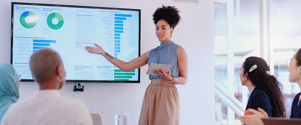 A person standing and pointing to a slide displayed on a large monitor, showing charts and graphs. They are holding a tablet and appear to be explaining the data to a group of seated people in a well-lit meeting room.