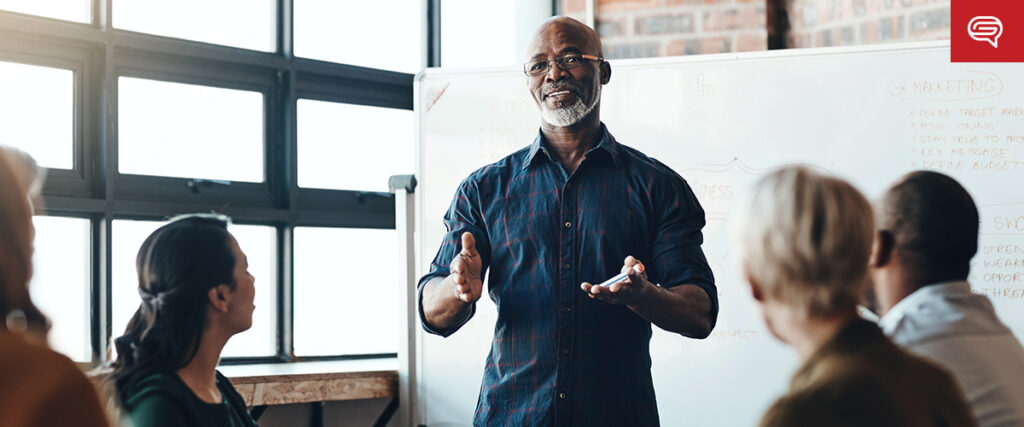 A man stands in front of a group of people, gesturing as he speaks. He is wearing glasses and a dark shirt. Behind him, a whiteboard with writing and a PowerPoint slide template are visible. The group appears to be in a brightly lit room with large windows.