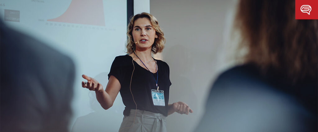 A woman with blonde hair dressed in a black shirt and gray pants is giving a presentation. She is gesturing with her hand while speaking into a headset microphone. A projection screen displaying a slide with a graph is in the background, with blurred figures in the foreground.