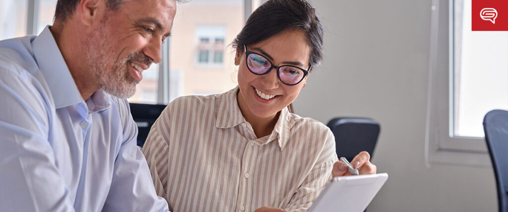 A man and a woman, both wearing glasses and business attire, sit next to each other in an office setting. The woman is holding and pointing at a tablet, smiling as they discuss the pitch deck displayed on the screen. The man is also smiling, engaged in conversation.