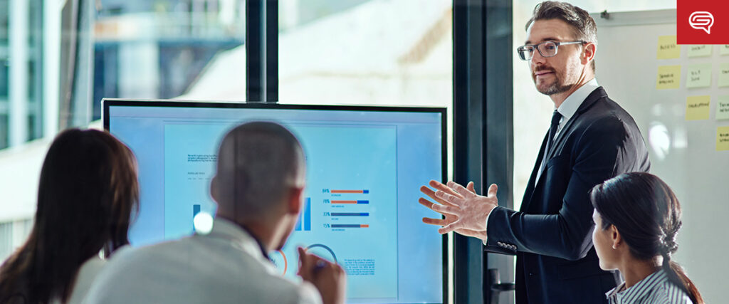 A man in a suit and glasses stands beside a large monitor, gesturing as he presents a PowerPoint slide to three colleagues seated around a table in a modern office. Sticky notes are visible on a glass wall in the background.