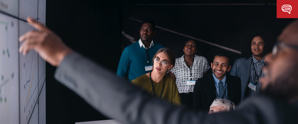 A group of people, mixed in gender and ethnicity, are gathered in a dark room, attentively looking at a slide being pointed to by a person in the foreground. Everyone is wearing business casual attire and name tags, suggesting a professional setting.