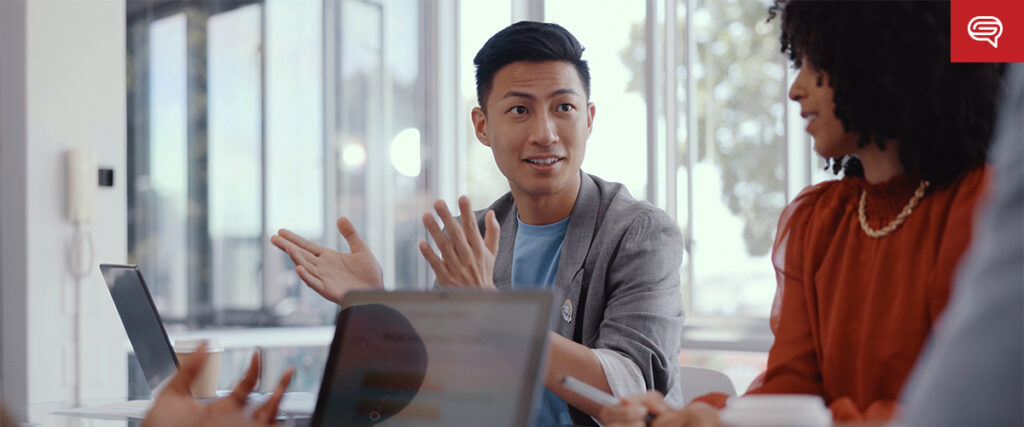 A man gestures while speaking during a meeting. He is seated at a table with laptops and a coffee cup, giving a presentation. A woman with curly hair sits to his right, listening attentively. Bright windows in the background light the modern office space.