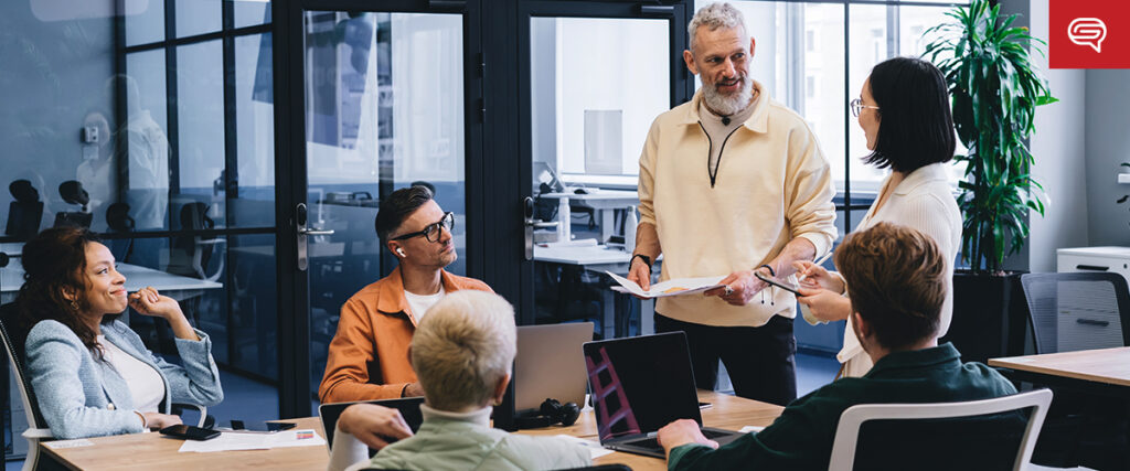 A group of six colleagues in a modern office, actively engaging in a discussion. One person stands holding papers while others sit with laptops and notebooks, going through their pitch deck. Large windows and glass partitions create a bright, open work environment.