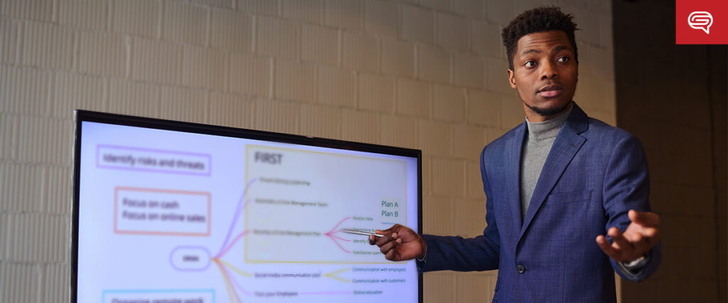 A man wearing a blue suit is standing and pointing at a PowerPoint slide displaying a flowchart during a presentation. The flowchart includes various colored lines connecting different text boxes. The background features a white wall with a speech bubble logo in the top right.