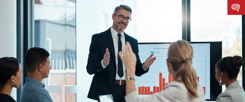 A man in a suit and tie presents a pitch deck displayed on a screen to a group of four seated colleagues in a modern office setting. One of the colleagues raises their hand, presumably asking a question or making a point. A large window reveals an urban backdrop.