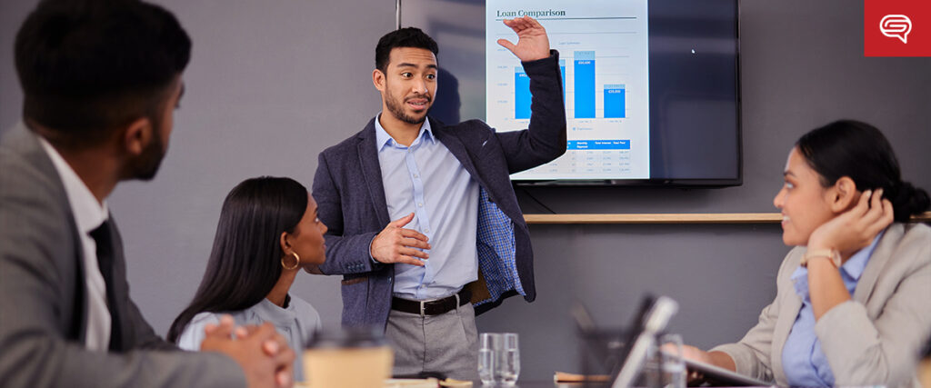 A man in a business suit is giving a presentation to three colleagues seated around a table. He is gesturing with one hand towards a PowerPoint slide displaying a chart titled "Loan Comparison." The atmosphere appears professional and focused.