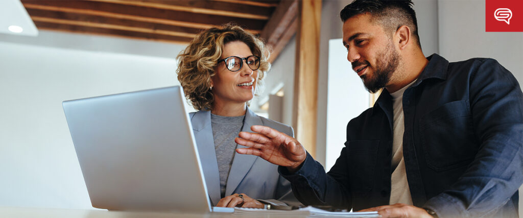 A man and a woman are sitting at a desk, engaged in a conversation. The woman is wearing glasses and a blazer, and the man is gesturing with his hand while explaining something. They both smile as they look at a PowerPoint presentation on the laptop. A speech bubble icon is in the top-right corner.