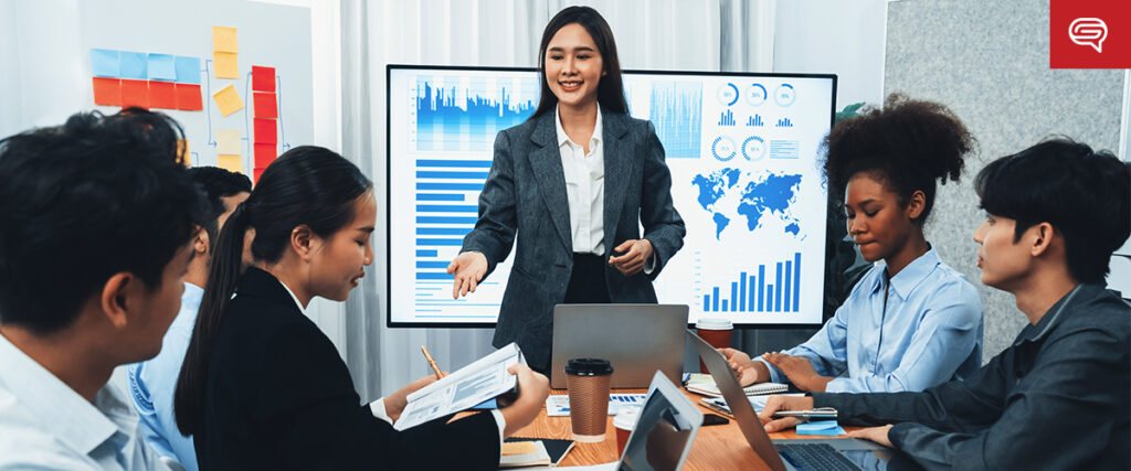 A woman in a gray blazer stands in front of a screen displaying various charts and graphs, including a world map. She is giving a presentation to a group of five seated individuals who are engaged and looking at papers and laptops.