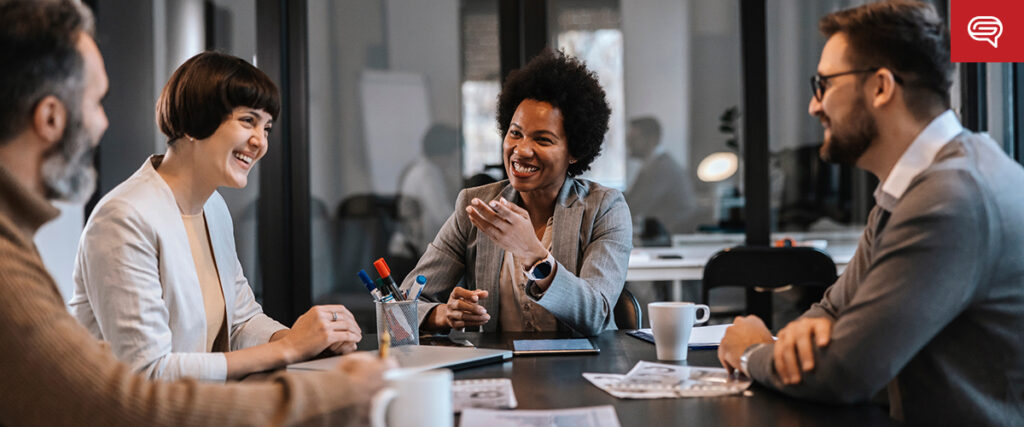 Four people are sitting around a table in an office setting, engaged in a lively discussion. They all appear to be smiling and laughing, suggesting a positive and collaborative atmosphere. The table has papers, cups, and pens on it, possibly as they review a pitch deck.