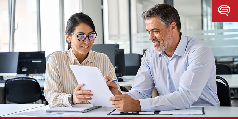 A man and a woman sit together at a desk in a bright office, reviewing a pitch deck. The man, with gray hair and beard, wears a light blue shirt, while the woman, with glasses and dark hair, is dressed in business attire. They both look engaged and smiling.