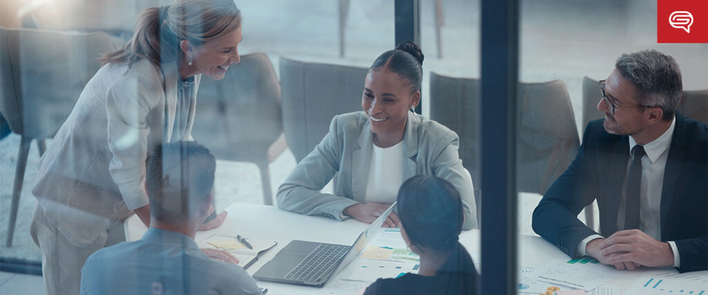 A diverse group of five business professionals engage in a meeting around a conference table. They are smiling and appear to be discussing a pitch deck and charts spread out on the table. In the background, glass walls provide a view of an office setting.