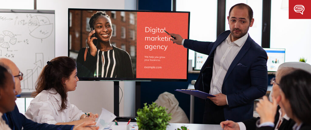 A business meeting with six people around a table. One man is standing and pointing to a presentation screen that displays a slide with a woman's photo, the text "Digital marketing agency," and a website link. The others are seated, listening attentively to the pitch deck.