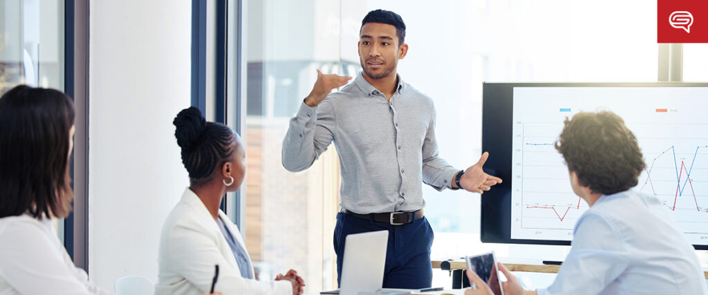 A man stands and gestures while presenting a PowerPoint to three seated colleagues in a modern office setting. A screen behind him displays a line graph. The group appears to be engaged in a discussion or meeting.