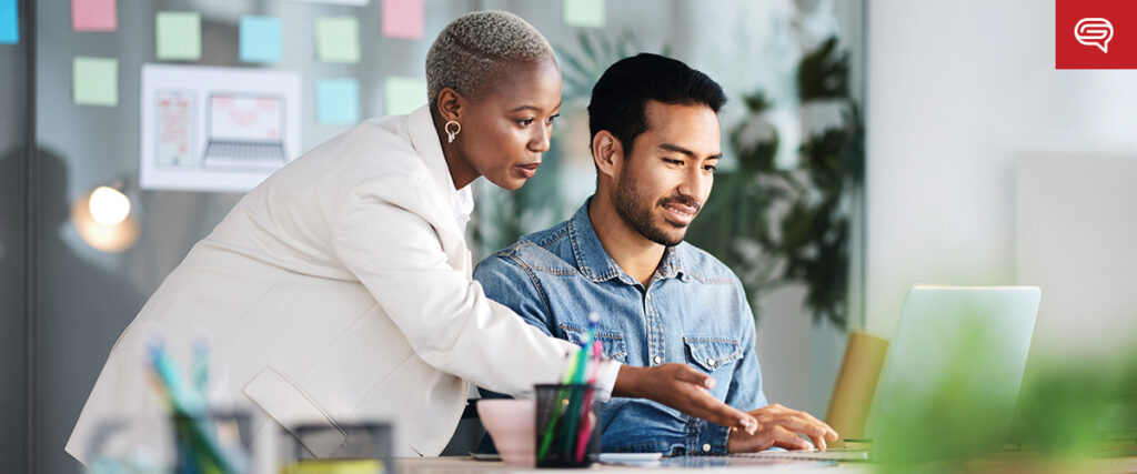 A woman in a white blazer stands beside a man in a denim shirt who is seated at a desk with a laptop. The woman points at the laptop screen, as they both focus on what seems to be their presentation. The background has sticky notes and office supplies, indicating an office setting.