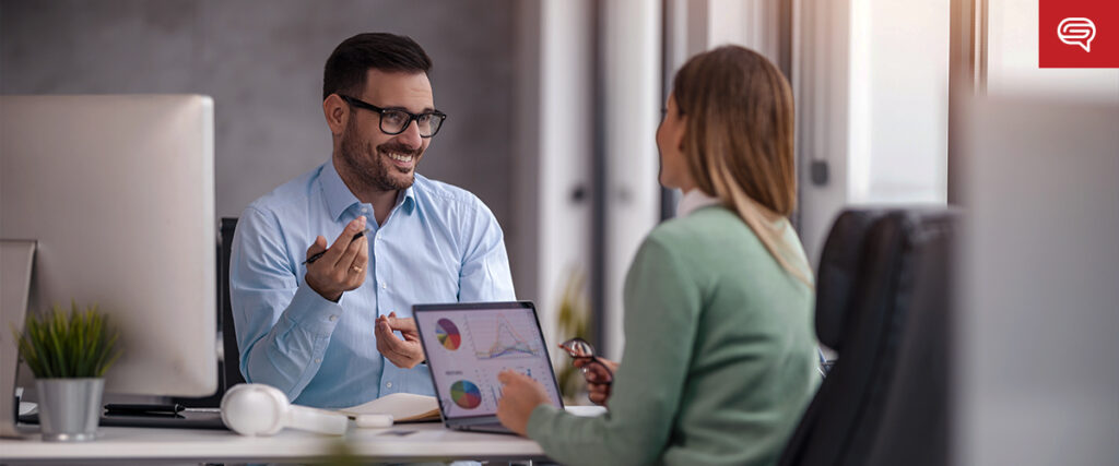 Two colleagues in a modern office having a discussion. A man in glasses and a light blue shirt smiles, holding a pen, while a woman with long hair in a green sweater points at a tablet displaying charts. The casual yet professional environment enhances their collaboration on the presentation.