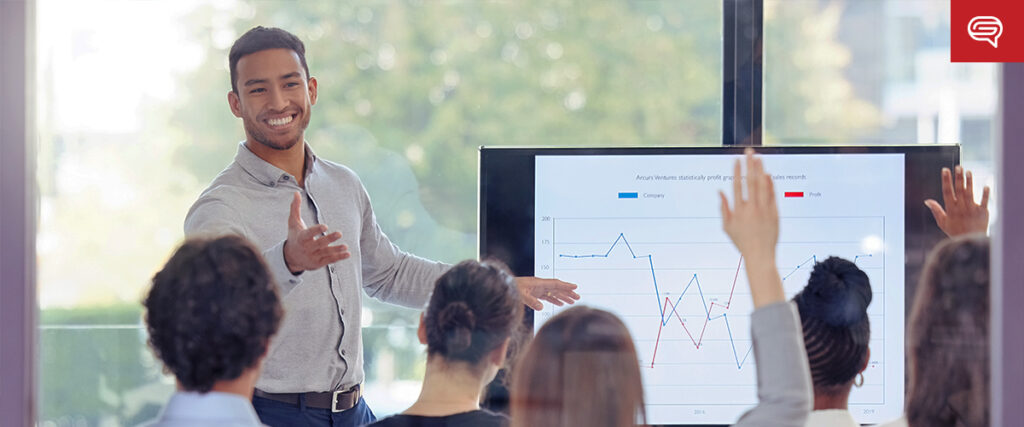 A man in a light blue shirt stands at the front of a room, smiling and gesturing towards a presentation screen displaying graphs. Several people are seated facing him, with some raising their hands. The background shows a window with greenery outside as he navigates through his PowerPoint slides.