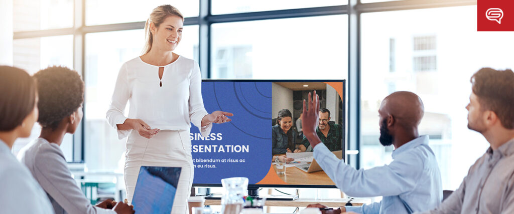 A woman stands and smiles while giving a presentation in a bright office. A screen behind her displays "Business Presentation" along with a slide of two colleagues. Four colleagues are seated around a table, one of whom is raising his hand to ask a question.