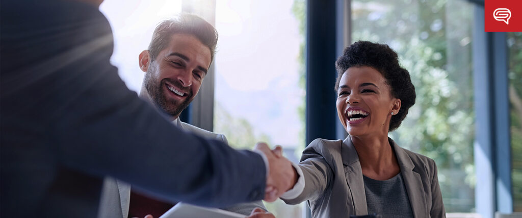 A woman and a man in professional attire are smiling and laughing as they shake hands with another person, whose back is to the camera. They appear to be in an office setting with large windows and natural light streaming in, perfect for capturing a moment to include in a PowerPoint presentation.