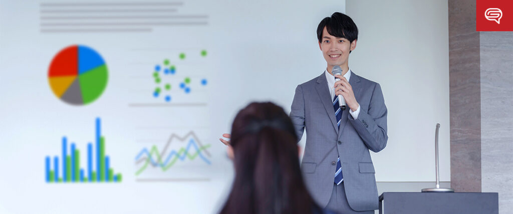 A person in a suit is giving a presentation using a microphone, pointing to a PowerPoint slide displaying various charts and graphs. An audience member's head is visible in the foreground, facing the presenter.