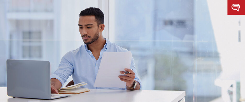 A man sits at a desk in an office, working on a laptop while holding a stack of documents and a pen. He is focused on his task, with an open book and slide template beside him. The background features large windows with a blurred cityscape view.
