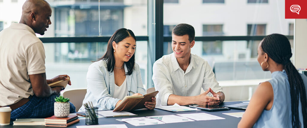 Four people are engaged in a meeting around a table with documents and a laptop displaying a PowerPoint slide. They appear to be discussing something important. One person sits on the edge of the table, while the others are seated in chairs. The background shows large windows with an urban view.