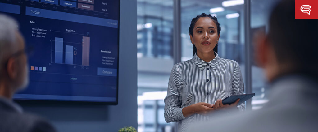 A professional woman stands holding a tablet, presenting a pitch deck in a modern office setting. Behind her, a large screen displays graphs and data. She is addressing two people sitting in front of her, whose heads are partially visible.