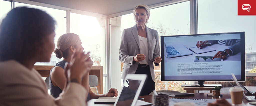 A person in a business suit stands and speaks in front of a TV displaying a slide template inside a bright office. Two other individuals are seated at the table, using laptops and taking notes. The setting suggests a professional meeting or presentation.