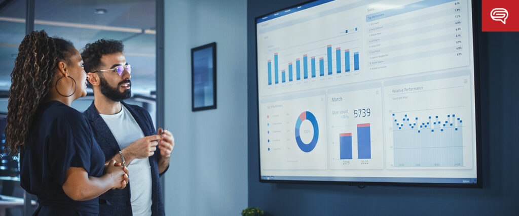 Two professionals stand in front of a large wall-mounted screen displaying various graphs and charts. The screen shows bar charts, pie charts, and line graphs with data points. The man is explaining something to the woman as they both analyze the information for their pitch deck.