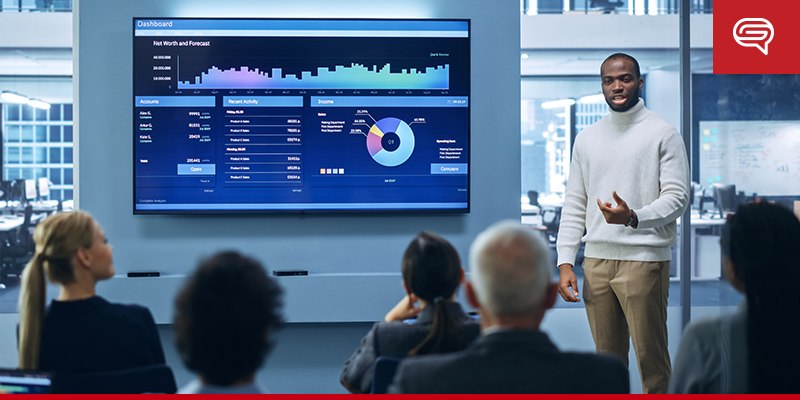 A man stands presenting data on a large screen to four seated colleagues in a modern office. The screen displays graphs and charts, and the room features glass walls with an urban view in the background. The scene depicts a business meeting with a well-designed slide template showcased during the presentation.