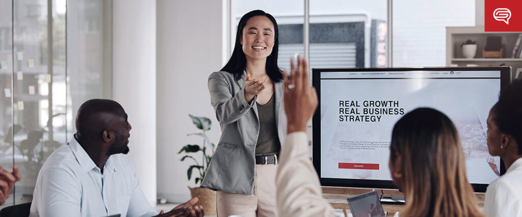 A woman stands and smiles while giving a presentation to a diverse group of people seated around a table. A large screen displays the text "Real Growth, Real Business Strategy" on a PowerPoint slide. Attendees are engaging, with one person raising their hand.