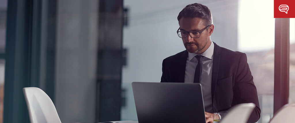 A man in a suit and tie sits at a desk in a modern office, working on a laptop. He is wearing glasses and appears focused on his screen, likely preparing a PowerPoint presentation. Sunlight filters through the large windows, creating a bright atmosphere. A small red icon is visible in the top right corner.