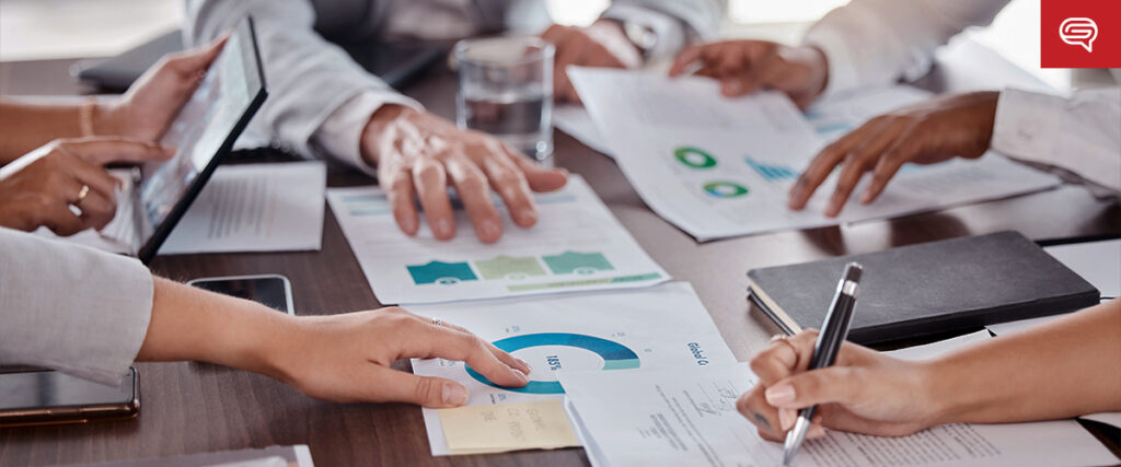 A group of people seated around a table engaged in a collaborative meeting. They are examining various charts, graphs, and documents. One person is using a tablet for the pitch deck, while another is writing notes with a pen. A glass of water and a notebook are also on the table.