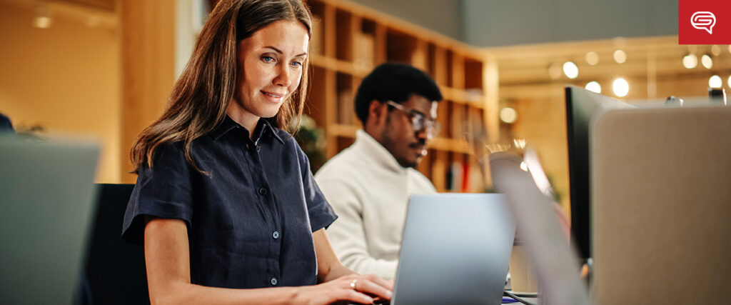 A woman and a man are working on laptops in a modern office space. The woman in the foreground is smiling and focused on her screen, likely preparing a presentation. The office has an open layout with shelves and warm lighting in the background.