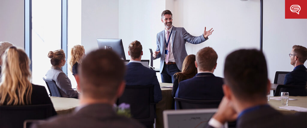 A man in a suit presents enthusiastically to a group of seated people in a conference room, using a pitch deck on the screen. He gestures with one hand while holding a microphone in the other. Attendees listen attentively, with laptops and documents on tables enhancing their focus.