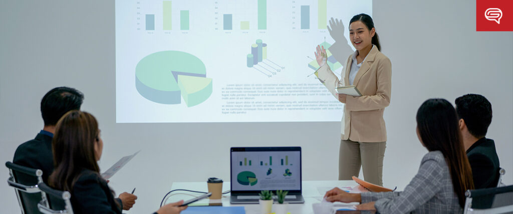 A woman in a beige blazer presents a slide template with data visualizations to a group of seated colleagues in a conference room. The projected slides show various charts and graphs, and a laptop on the table displays similar visuals. The setting appears professional and modern.
