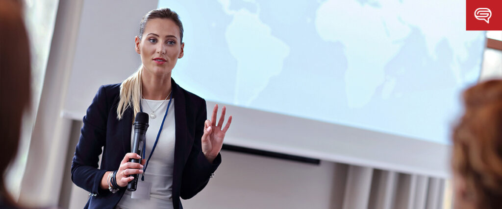 A woman in a business suit holds a microphone while giving a presentation. She stands in front of a projected world map slide template, addressing an audience. Her hand is raised, gesturing to emphasize a point.