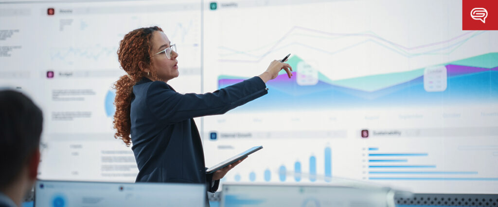 A woman wearing glasses and a dark blazer is pointing at a large screen displaying various charts and graphs. She holds a tablet in her other hand. The screen shows PowerPoint slides with data visualizations, including line graphs and bar charts, in a modern office setting.